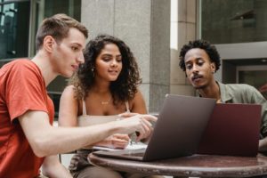 Group of diverse young male and female diverse colleagues discussing project details while working together on laptops in outdoors cafe